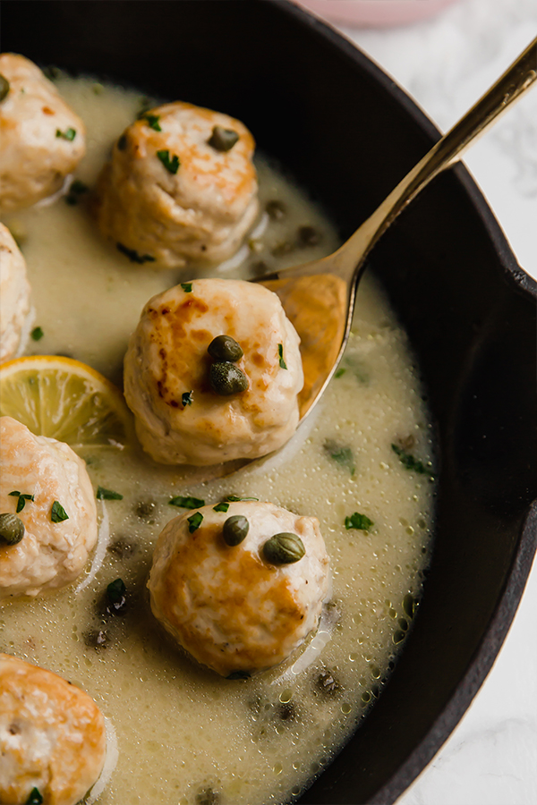 Chicken piccata meatballs in sauce in a pan with a spoon scooping out one meatball.