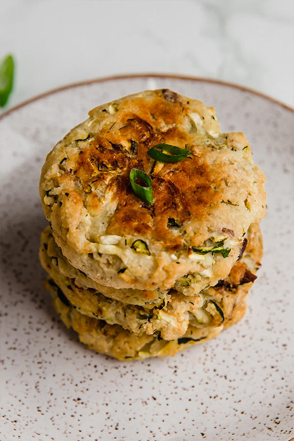 A stack of bacon zucchini breakfast patties lying on a plate with extra herbs.