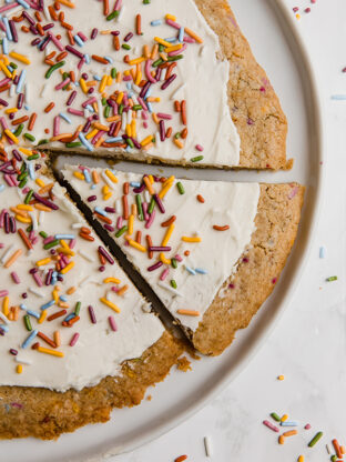 A white plate with a funfetti cookie pizza on it with a slice cut out but not yet removed.