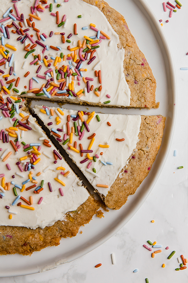 A white plate with a funfetti cookie pizza on it with a slice cut out but not yet removed.