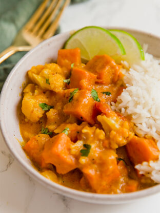 A white bowl filled with cauliflower rice and sweet potato cauliflower curry and fresh lime slices.