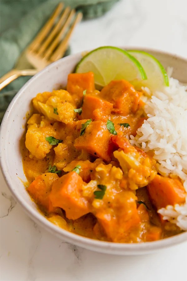 A white bowl filled with cauliflower rice and sweet potato cauliflower curry and fresh lime slices.