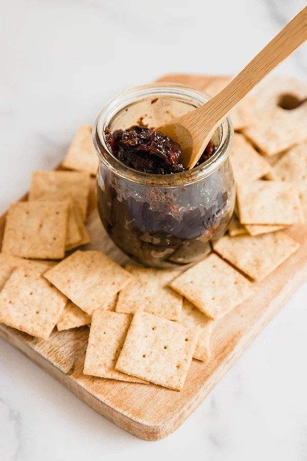 A wooden board filled with crackers and a glass jar with apple bacon jam and a wooden spoon in the jar of jam.
