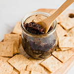 A wooden board filled with crackers and a glass jar with apple bacon jam and a wooden spoon in the jar of jam.