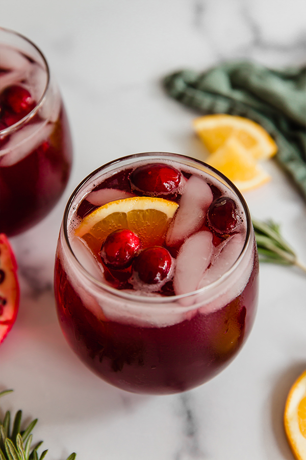 Pomegranate mocktail in a glass with ice, cranberries, and orange slices with rosemary and orange slices in the background.