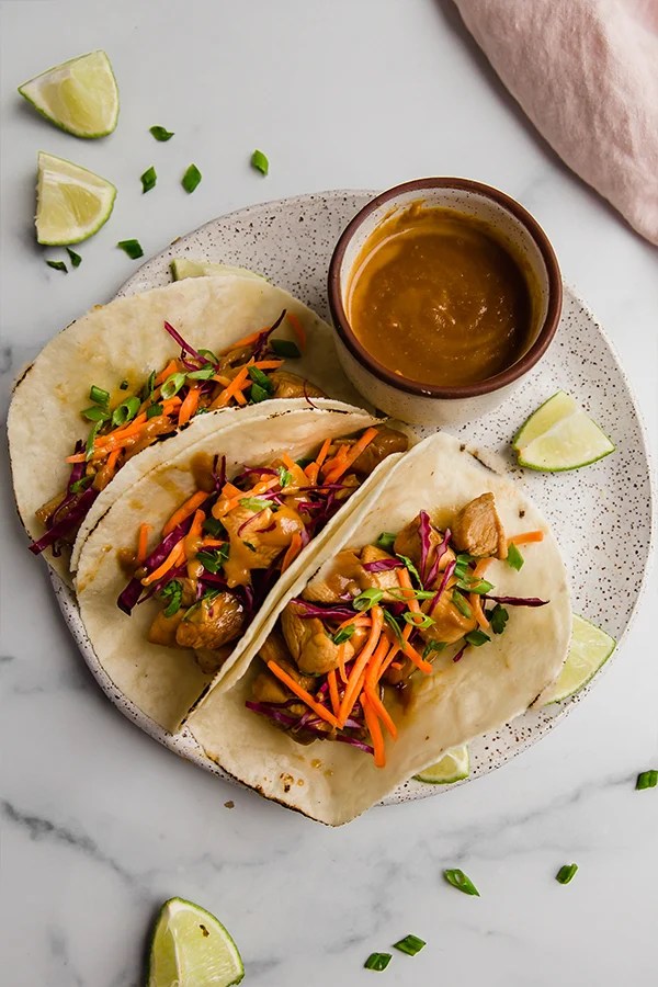 Three thai chicken tacos on a plate with sauce on the side.