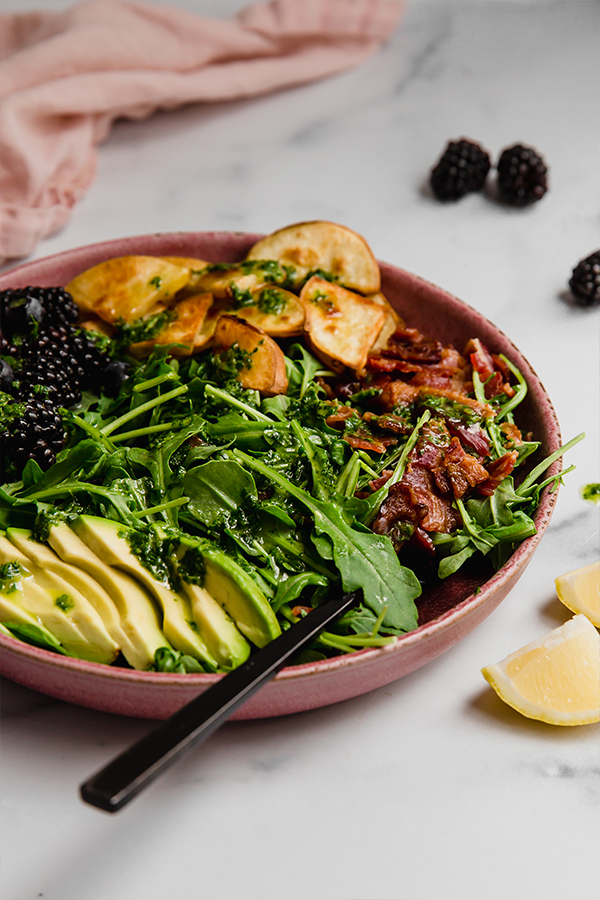 Breakfast salad topped with bacon, avocado, sweet potato, and berries served in a bowl with fork