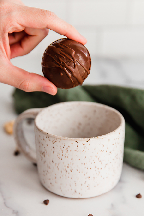 Person holding a hot cocoa bomb over a speckled mug.