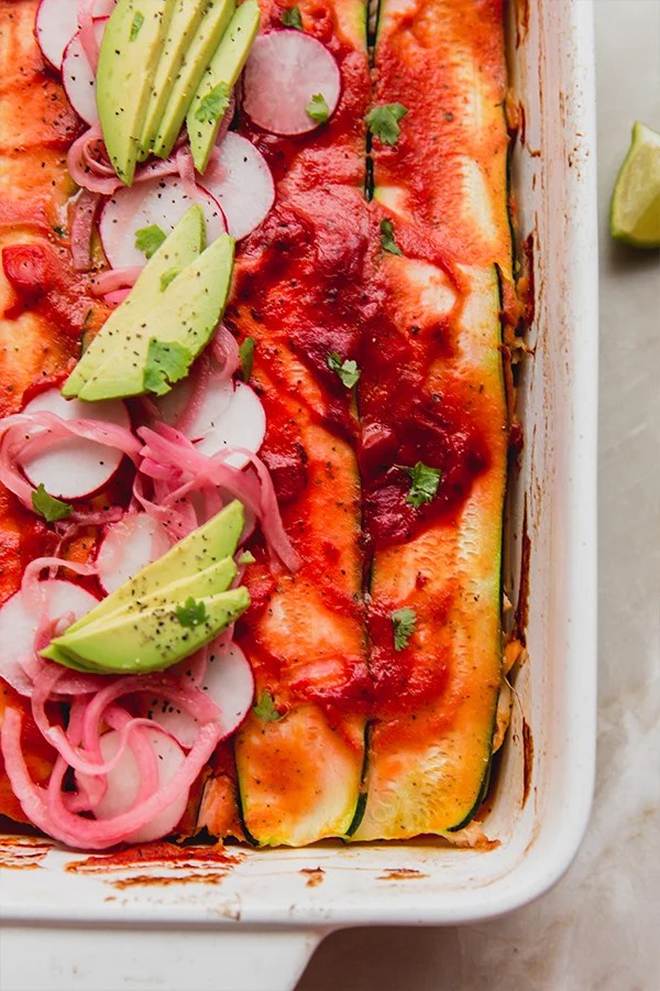 Zucchini enchilada casserole in a baking dish topped off with enchilada sauce, slices radish, avocado, pickled onion and cilantro.