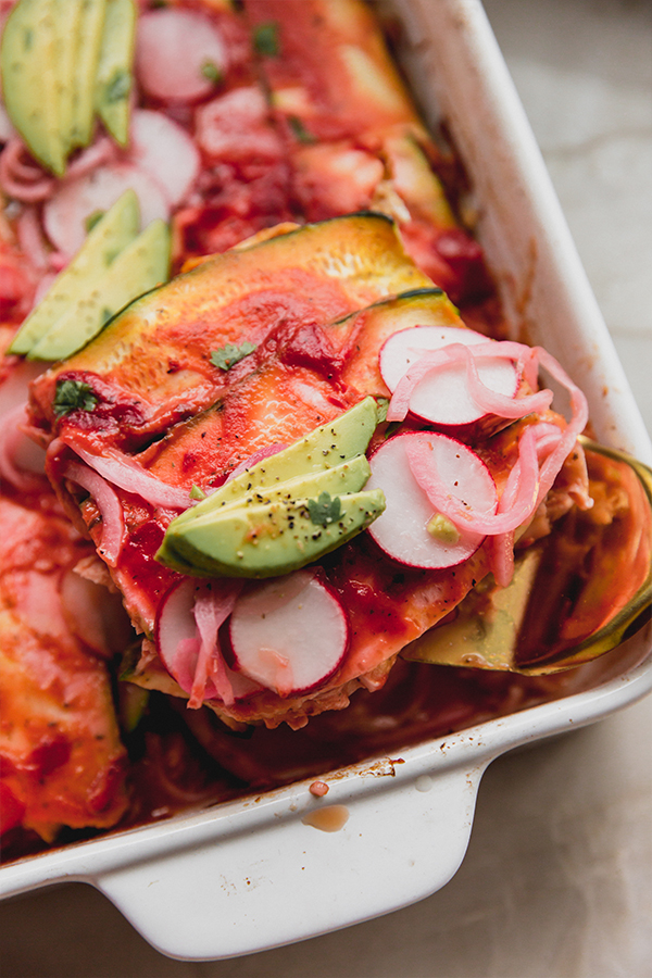 Zucchini enchilada casserole in a baking dish topped off with enchilada sauce, slices radish, avocado, pickled onion and cilantro, with a slice being taken out.