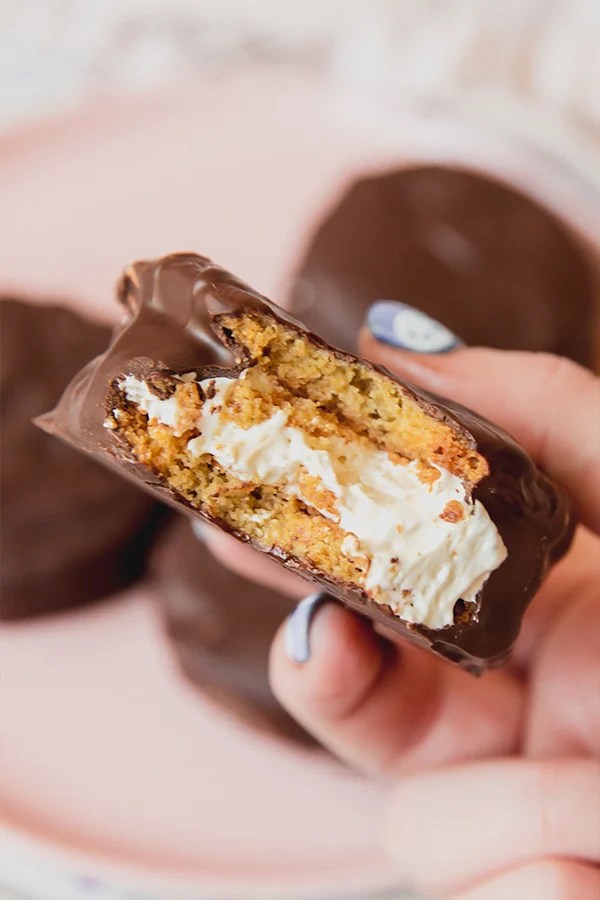 Chocolate coated pie being held with a hand, with a bite taken out of it to show the cookies sandwiching the marshmallow cream.