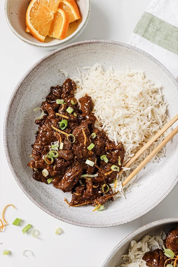 Orange beef topped with green onion and orange zest in a bowl with rice and chopsticks.