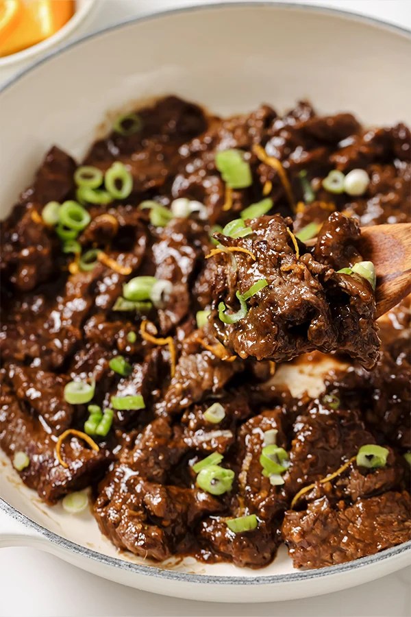 Orange beef topped with green onion and orange zest in a white enamel pot, with a wooden spoon scooping a serving.