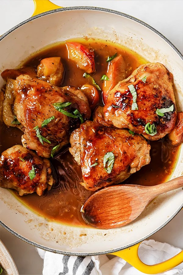 One pan balsamic peach chicken thighs topped with basil in a yellow pan with a wooden spoon.