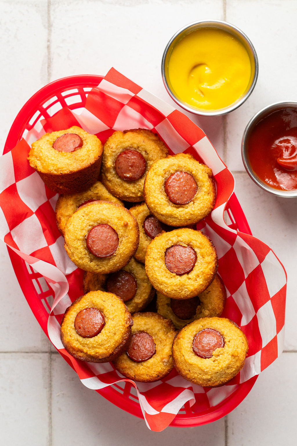 Corndog mini muffins in a red basket with checkered paper and a side of ketchup and mustard.