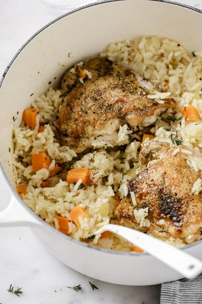 One pot baked chicken and rice in a large white pot, topped with fresh herbs, with a spoon dipping into the pot.