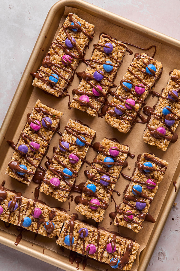 Kudos bars with candy on top and melted chocolate on a baking sheet, ready to be set.