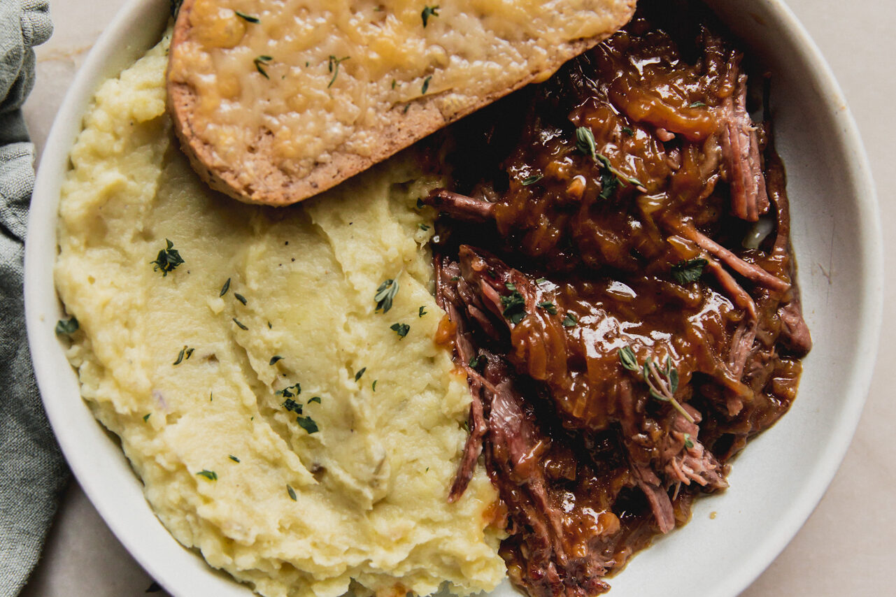 French onion pot roast in a shallow bowl with mashed sweet potato, cheese toast, and topped with herbs.