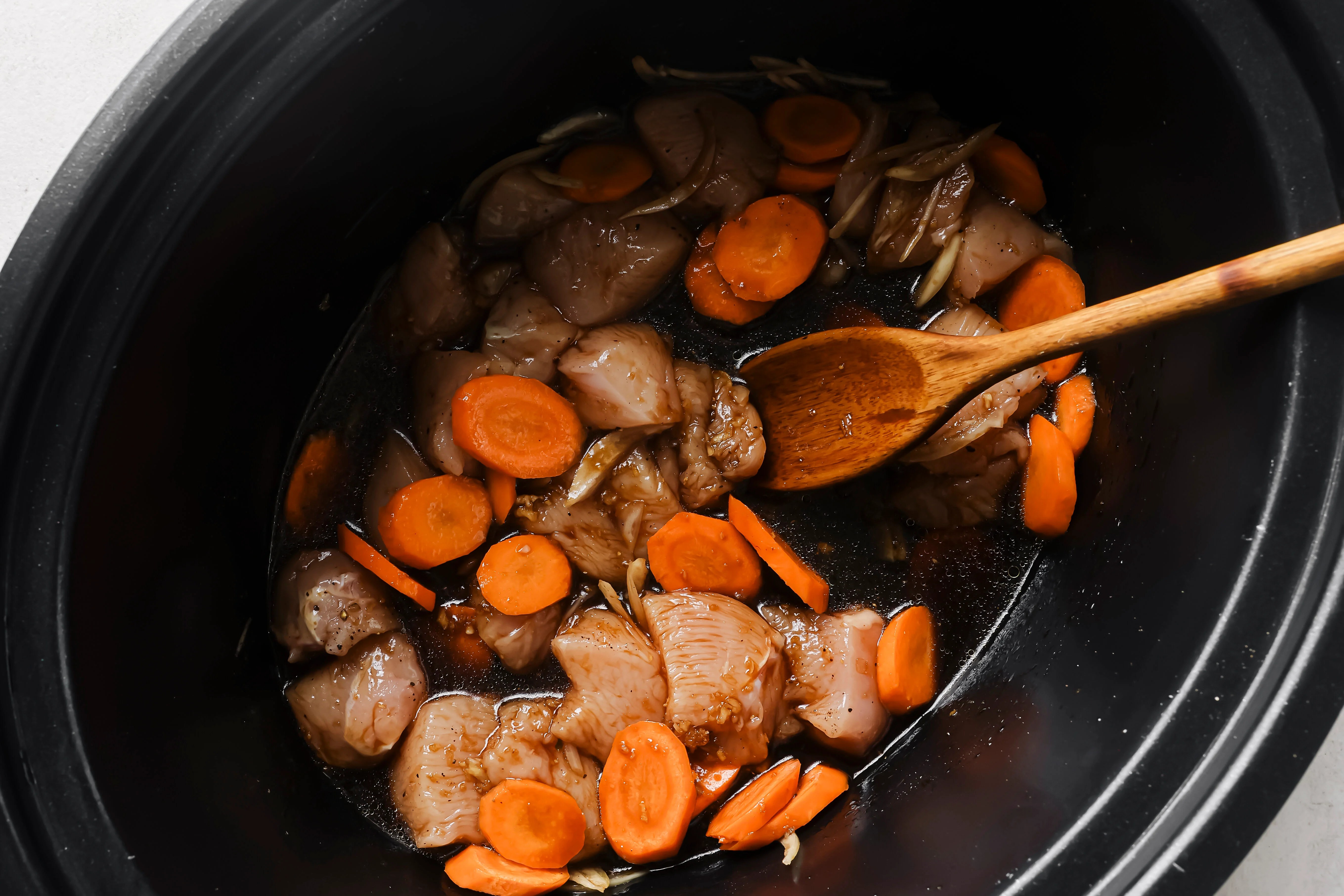 Chicken and broccoli uncooked in a slow cooker with the sauce and a wooden spoon.