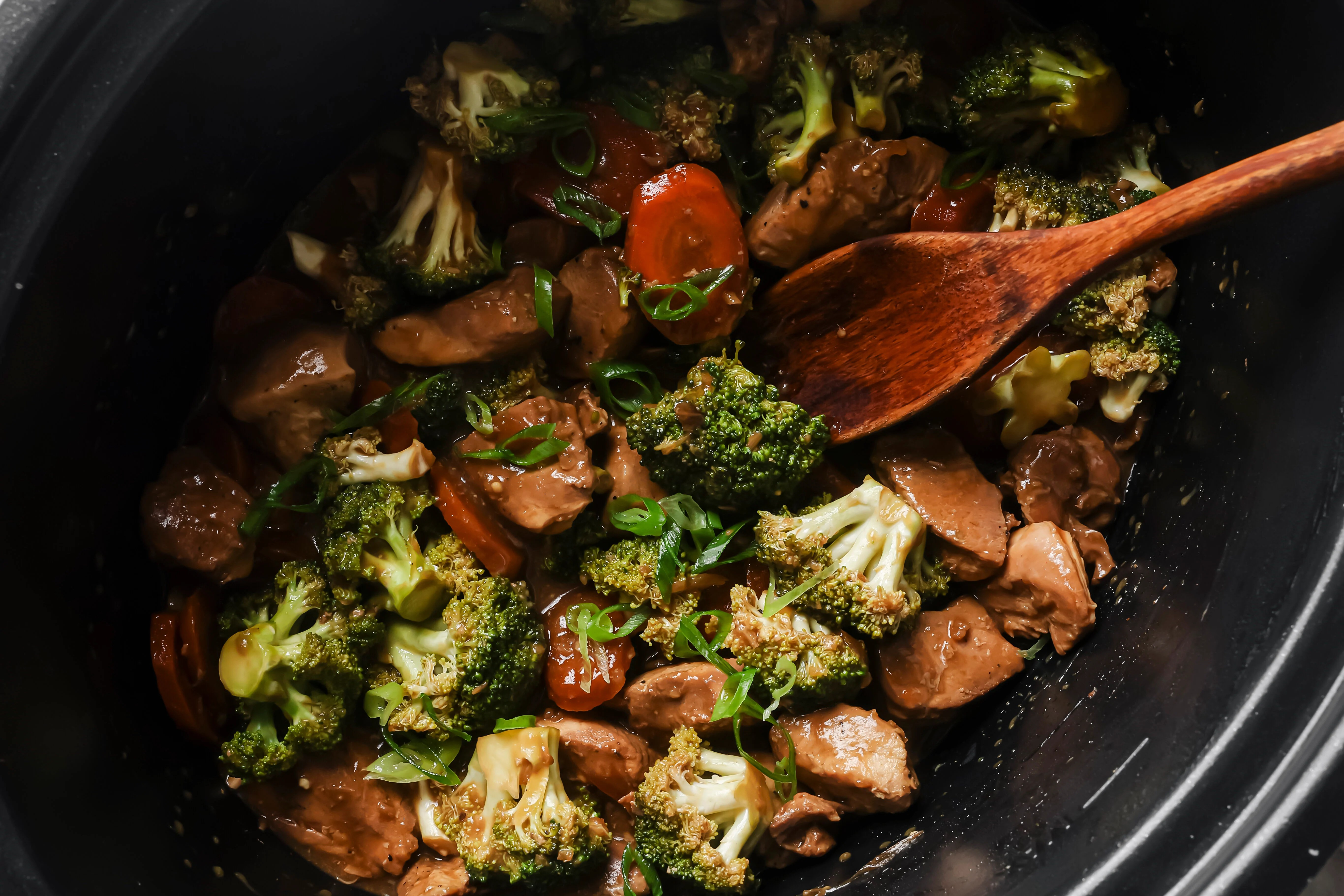 Fully cooked chicken and broccoli in a slow cooker topped with green onion, with a wooden spoon.