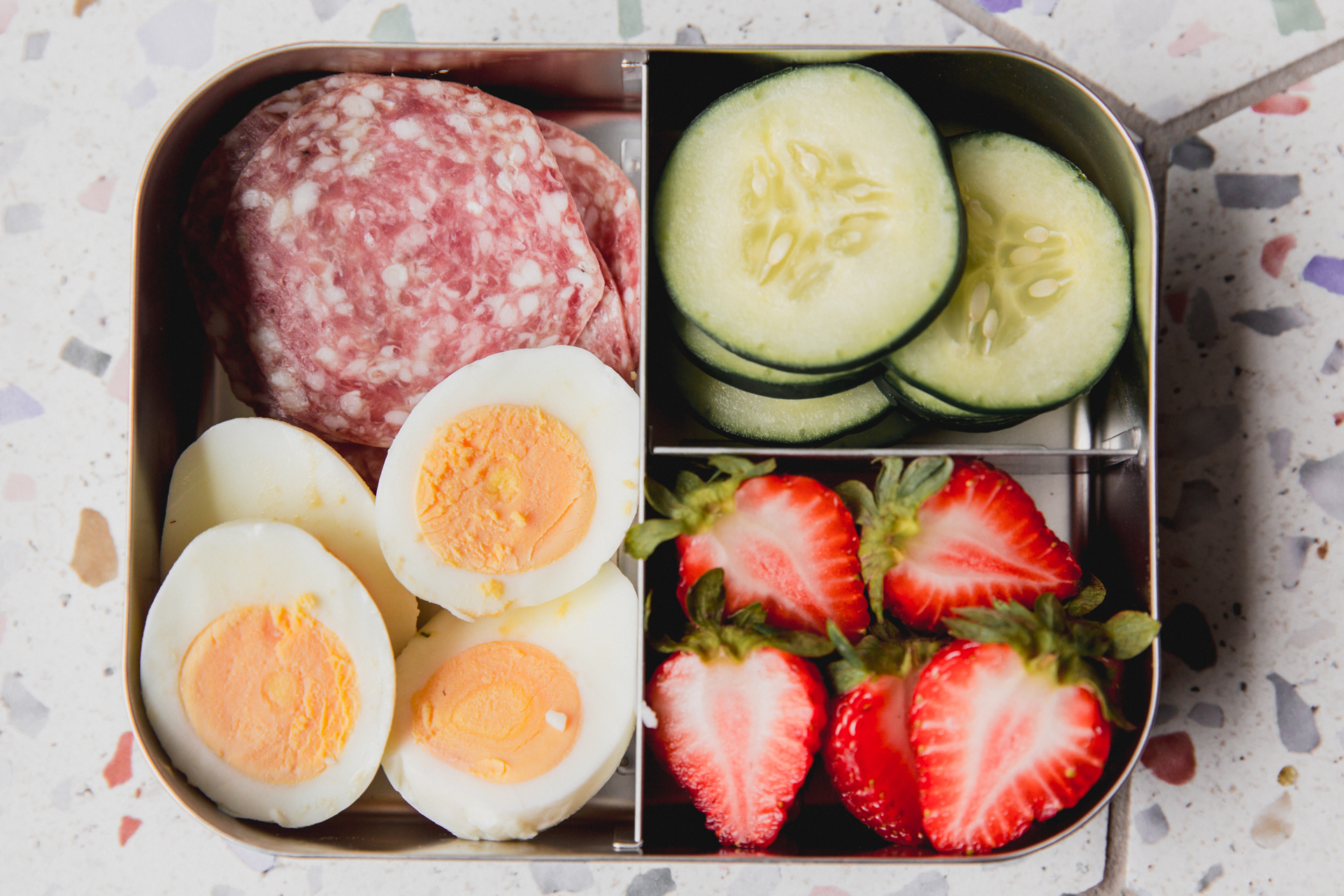 Divided lunch container with lunch meat, sliced boiled eggs, sliced cucumber, and sliced strawberries.