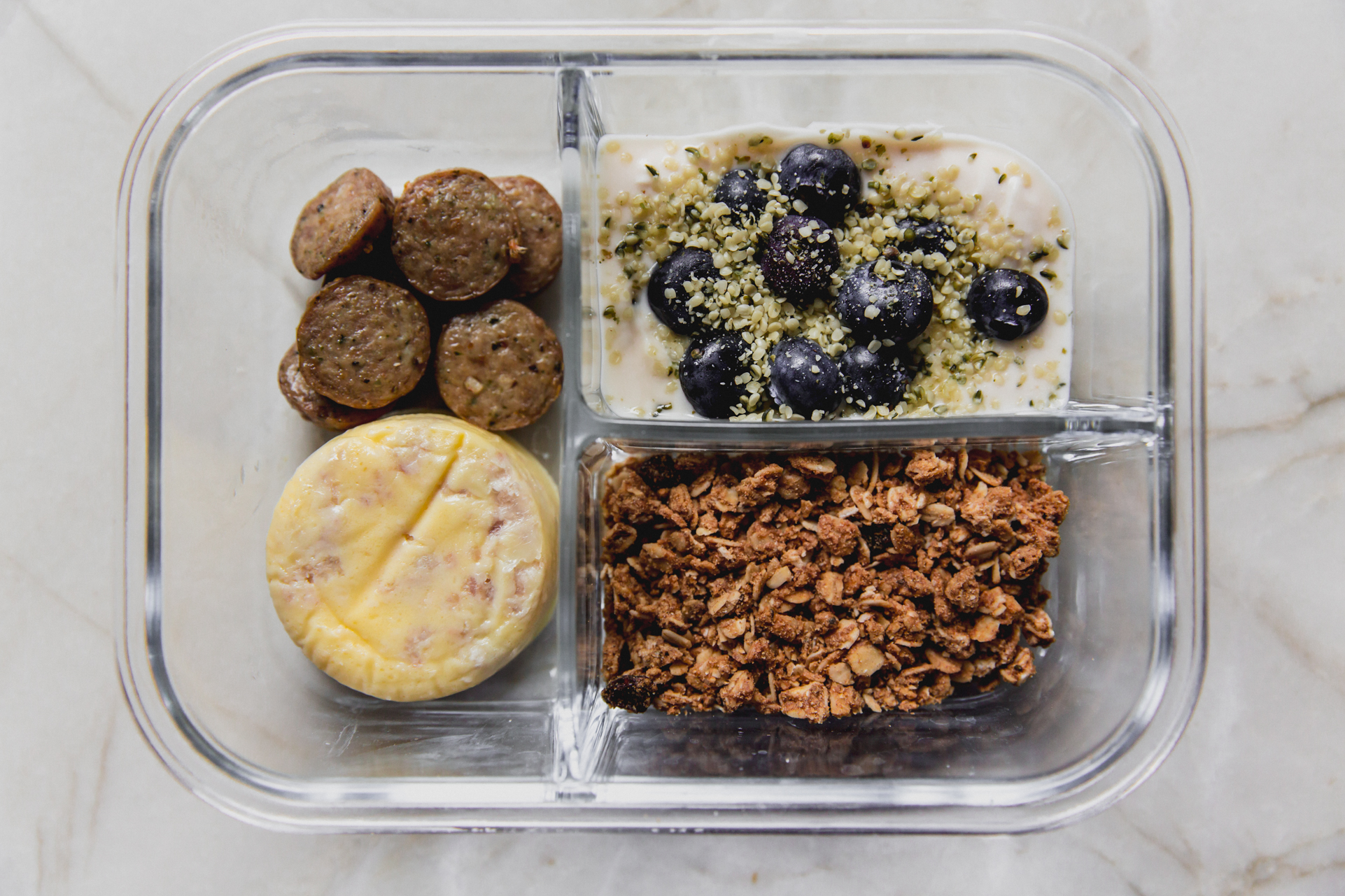 Clear divided lunch container with sausage, egg, granola, and yogurt.