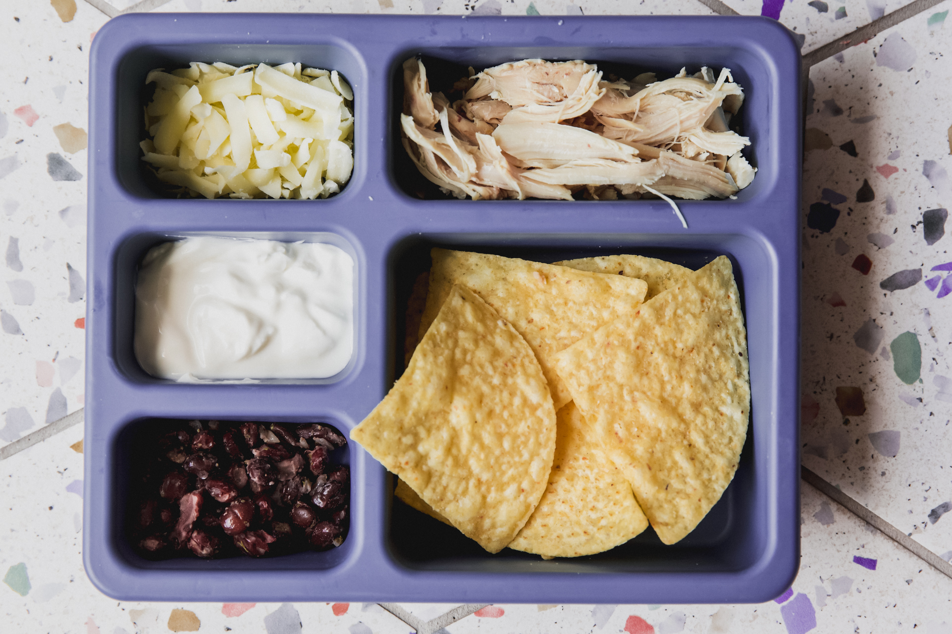 Divided lunch container with tortilla chips, shredded chicken, shredded cheese, cooked black beans and sour cream.