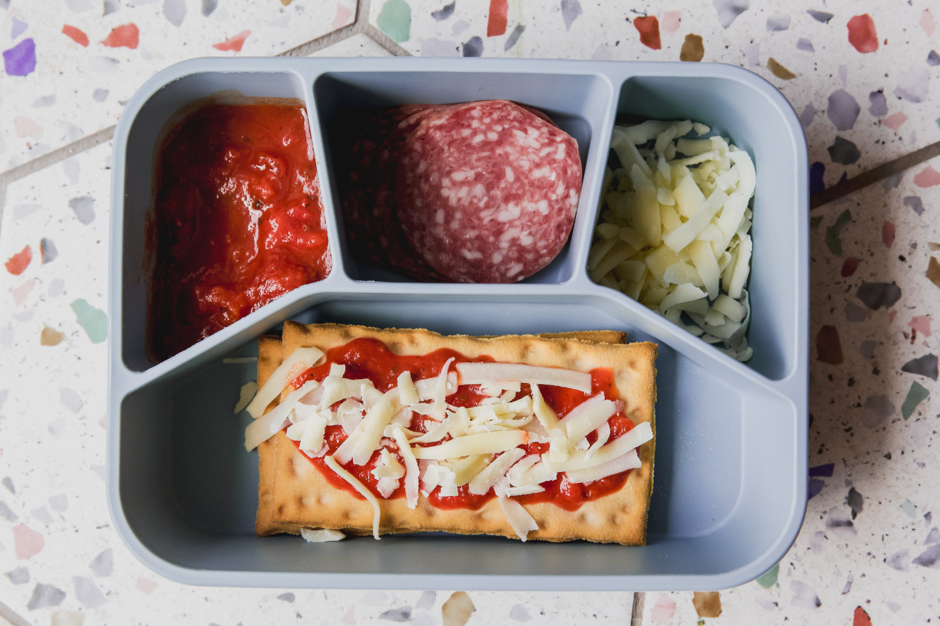 Blue lunch container with pizza sauce, meat, cheese, and a prepared flatbread pizza.
