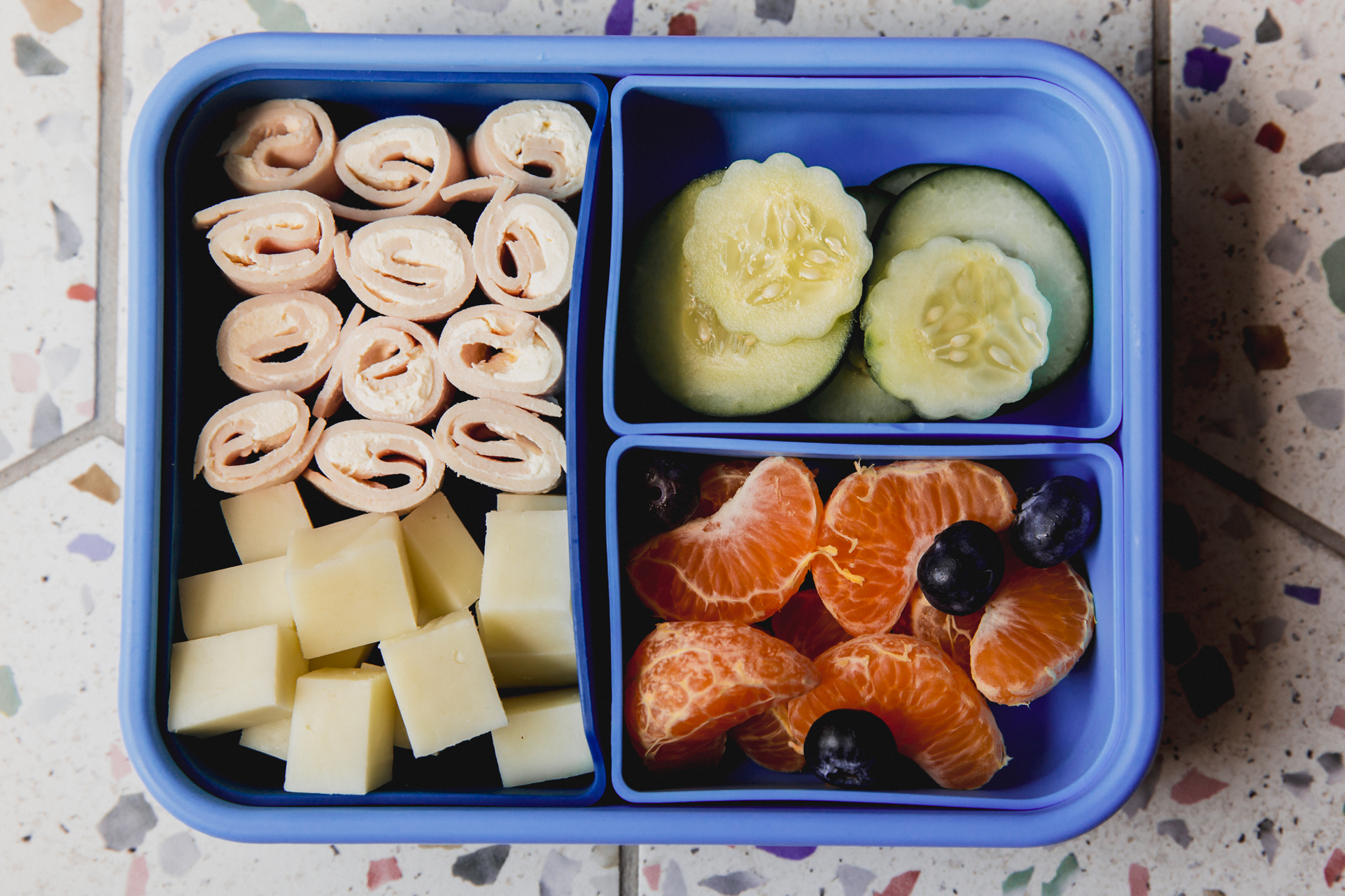 Blue divided lunch container with turkey roll ups, cubed cheese, sliced cucumber, clementines and blueberries.