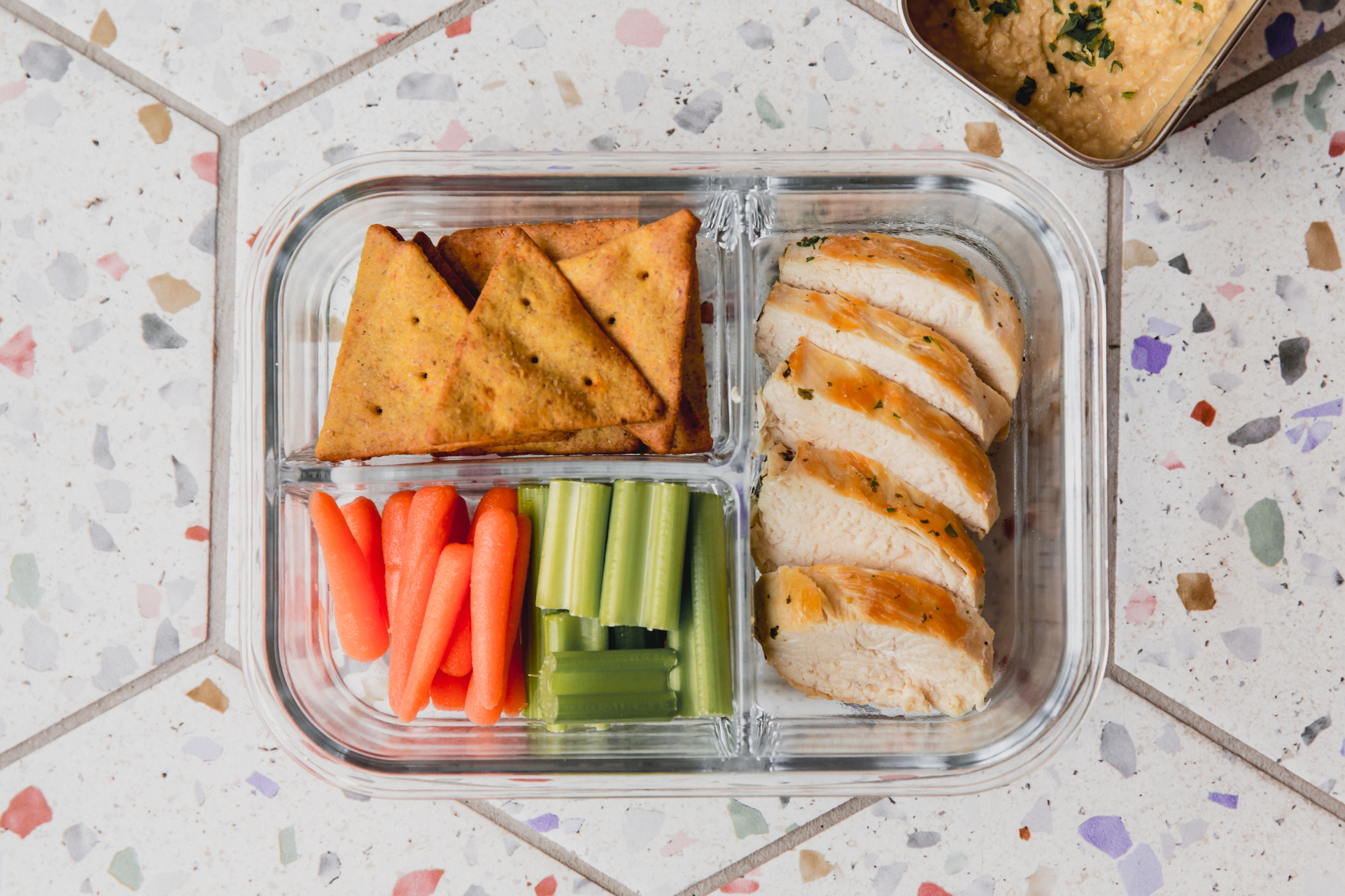 Clear divided lunch container with chicken, veggies, and pita chips.