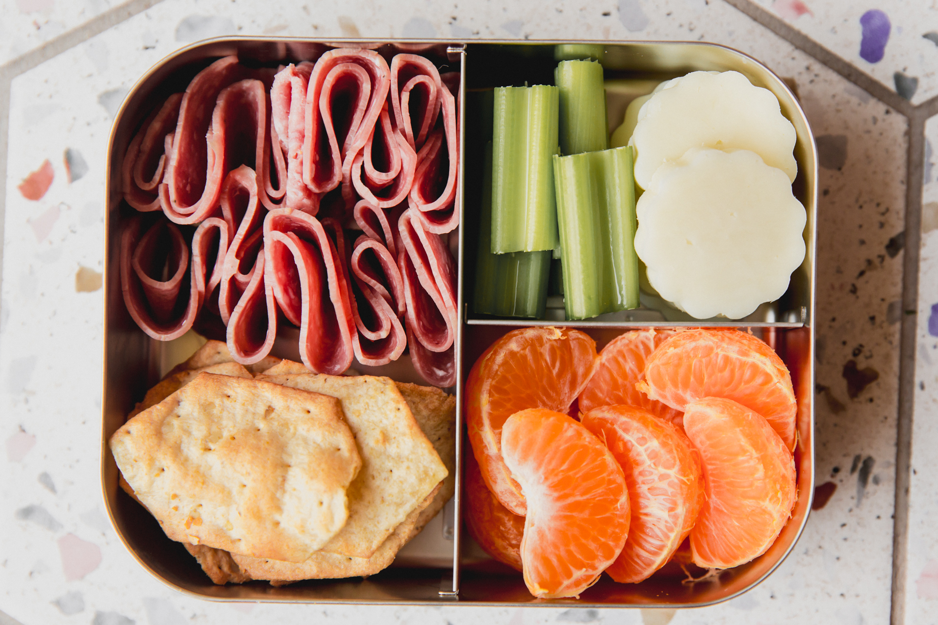 Stainless steel divided lunch container with cheese, celery, segmented clementine, lunch meat, and crackers.
