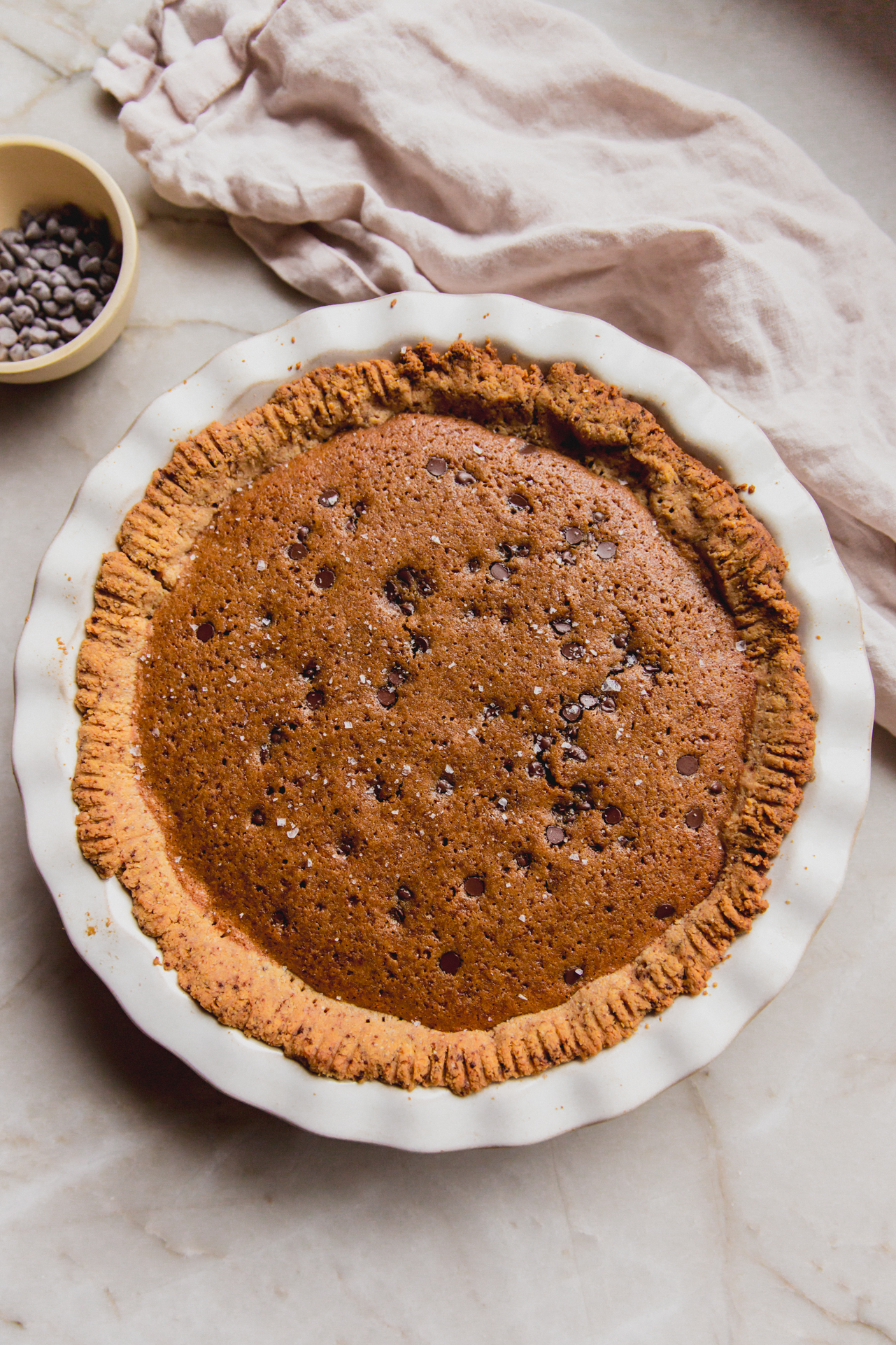Chocolate chip cookie pie in a white pie pan with a bowl of chocolate chips on the side. 