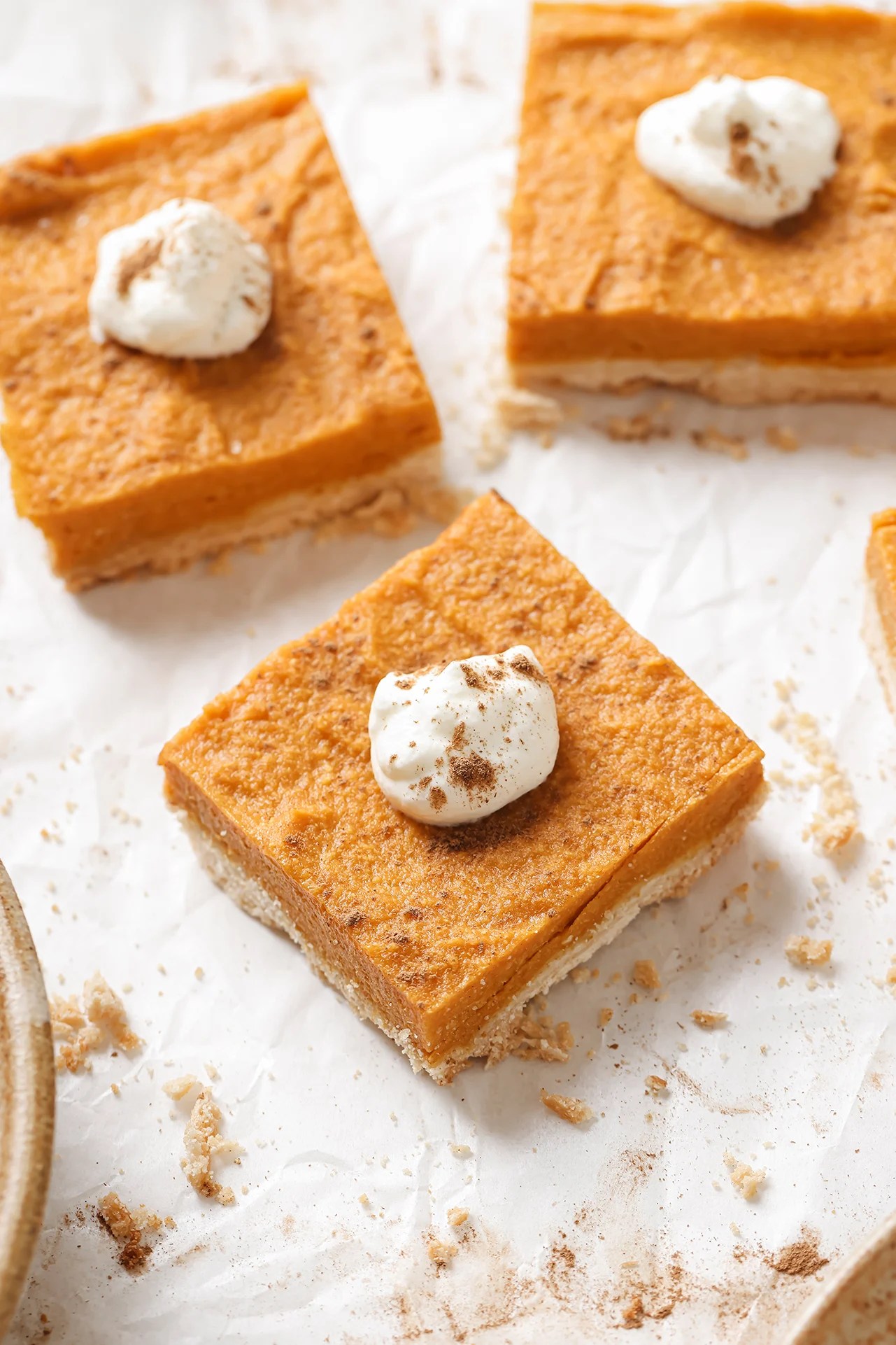 Three AIP pumpkin pie bars on parchment paper, topped with coconut cream and cinnamon.