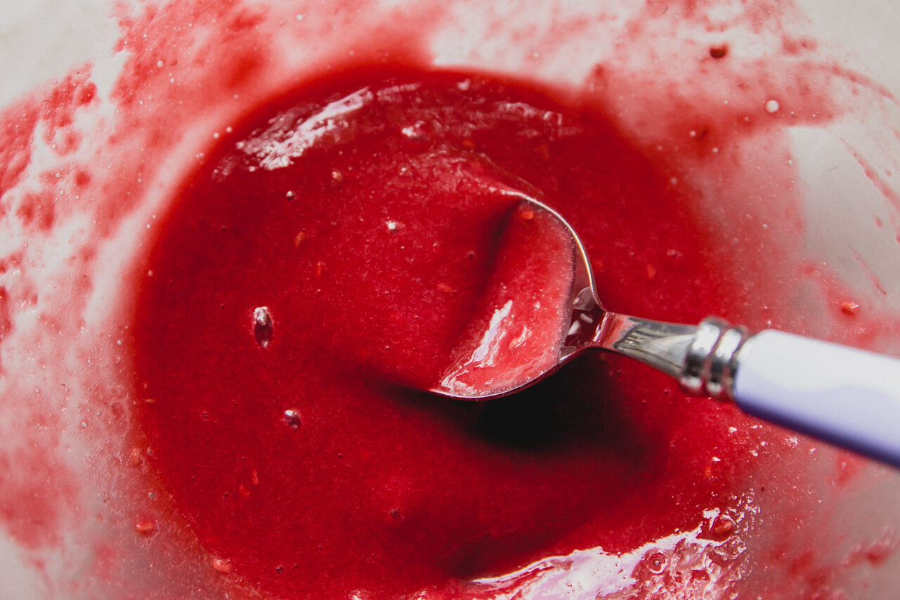 A glass bowl with raspberry mash being mixed by a spoon.