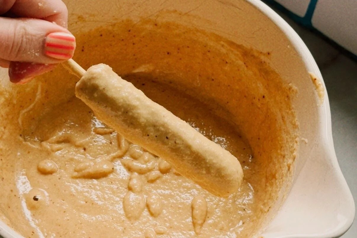 A white ceramic bowl filled with pancake batter with a hand dipping one chicken sausage skewer into it to coat with batter.