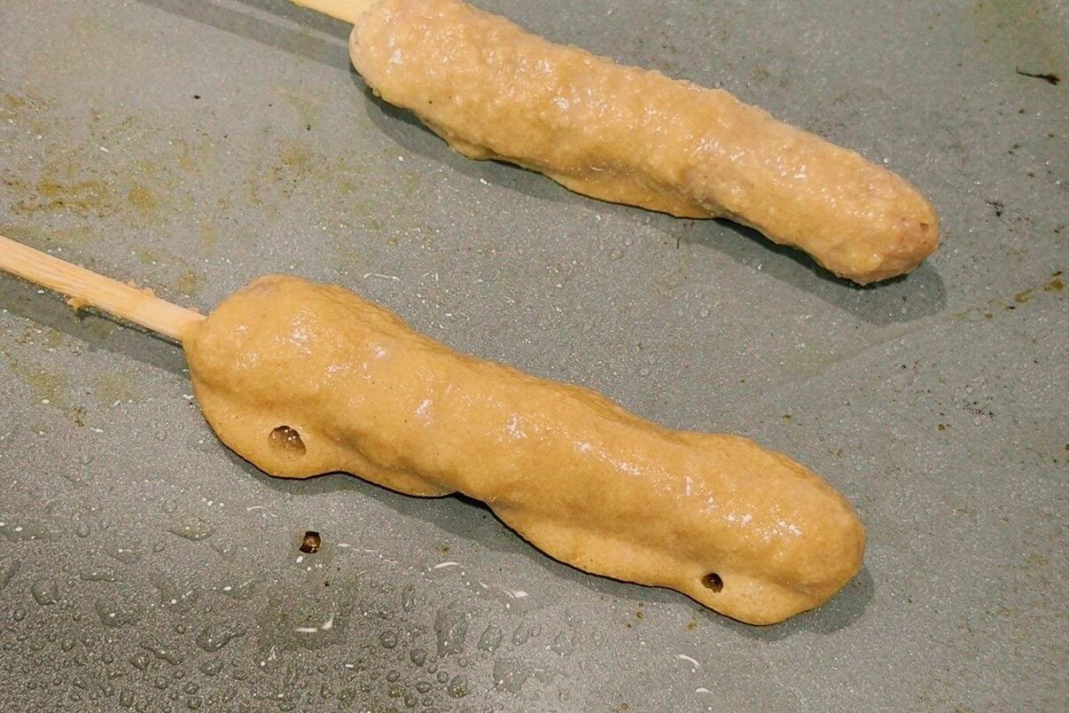 Two chicken sausage pancake skewers cooking on a nonstick pan.