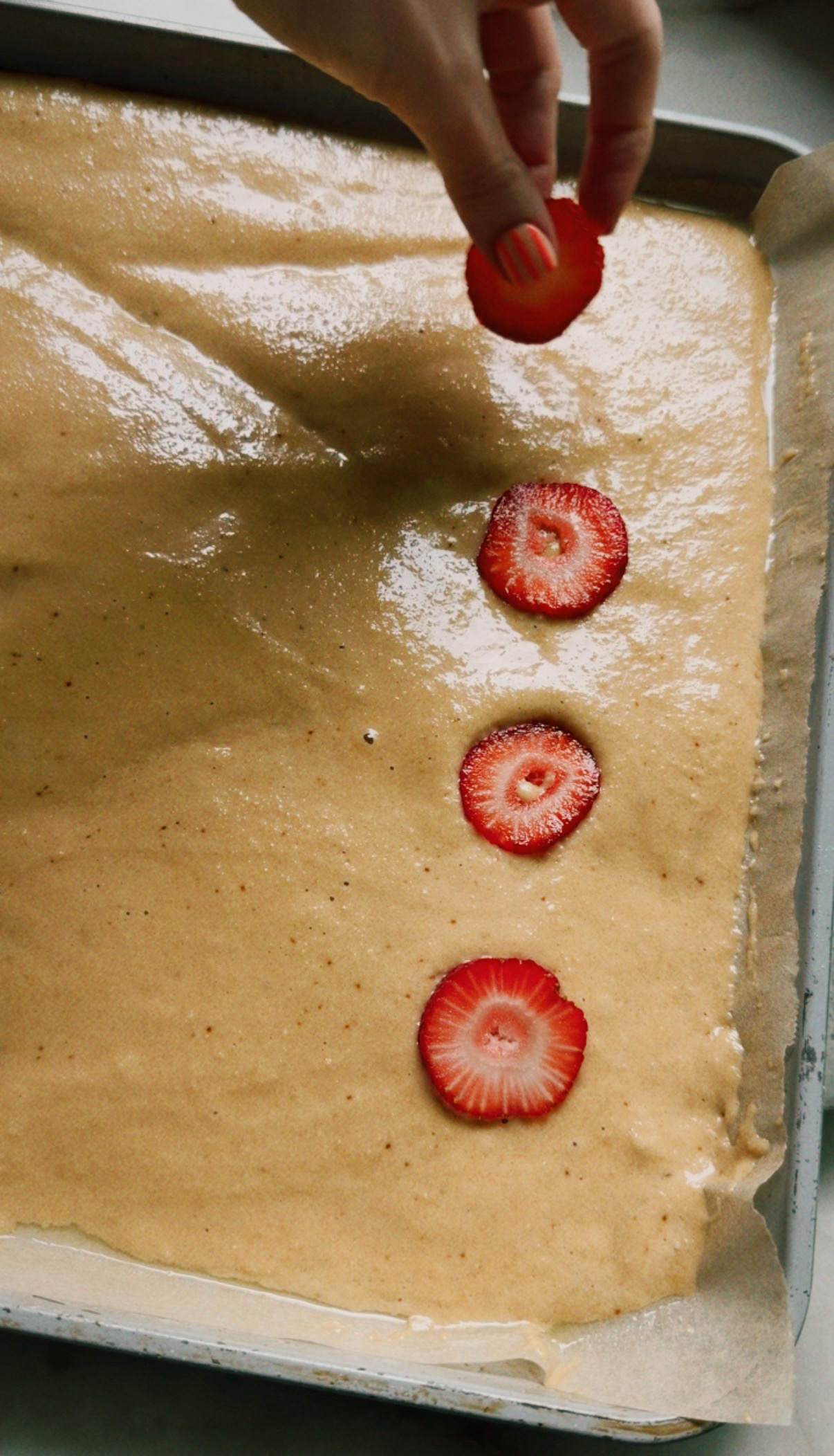 A hand placing sliced strawberries on unbaked sheet pan pancake batter