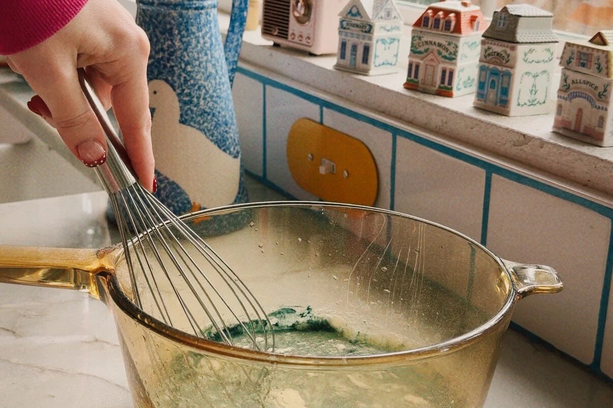 A glass bowl with apple juice, bue spirulina, and gelatin being mixed with a whisk.