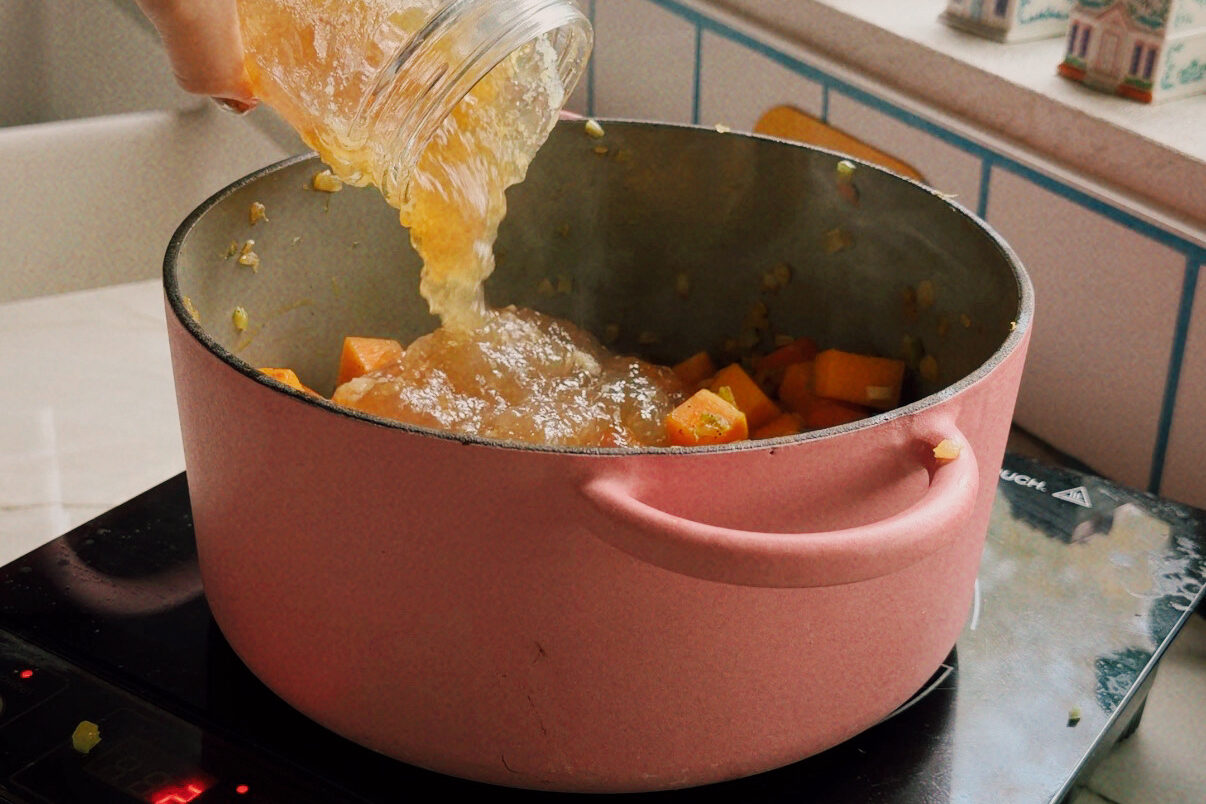 A glass jar of bone broth being poured into the pot of sauted veggies.
