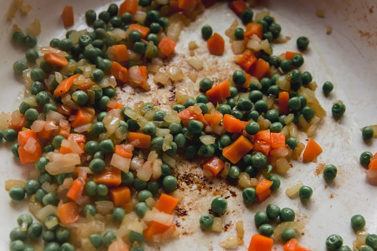 The onions, carrots, and peas sauting in a pan.