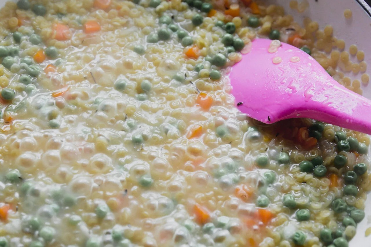 The pan filled with sauted veggies and broth with a pink spatula stirring.