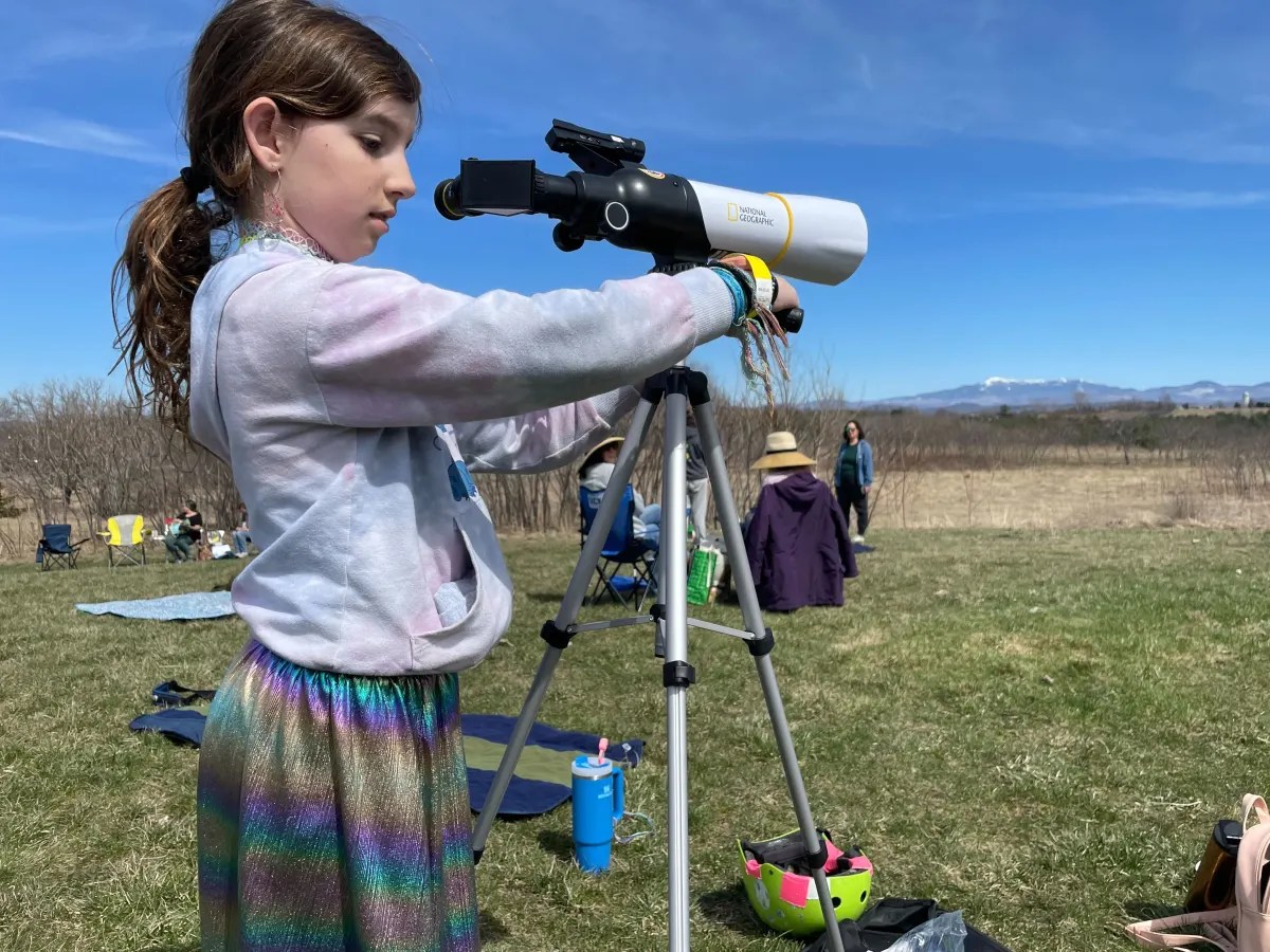 A young girl using a telescope outdoors on a sunny day.
