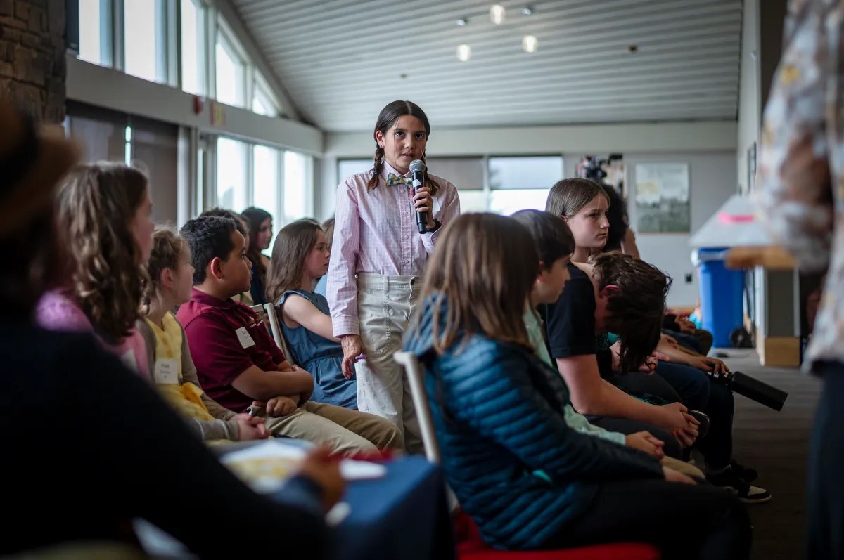 A person standing while speaking before a seated group in an indoor setting.