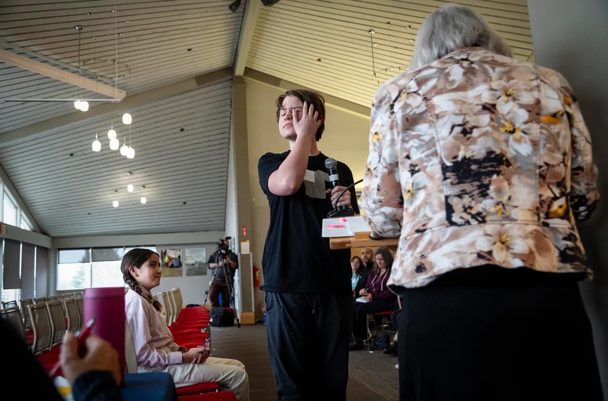 A person stands in a room expressing emotion with their hand on their face, as onlookers watch from seated positions.