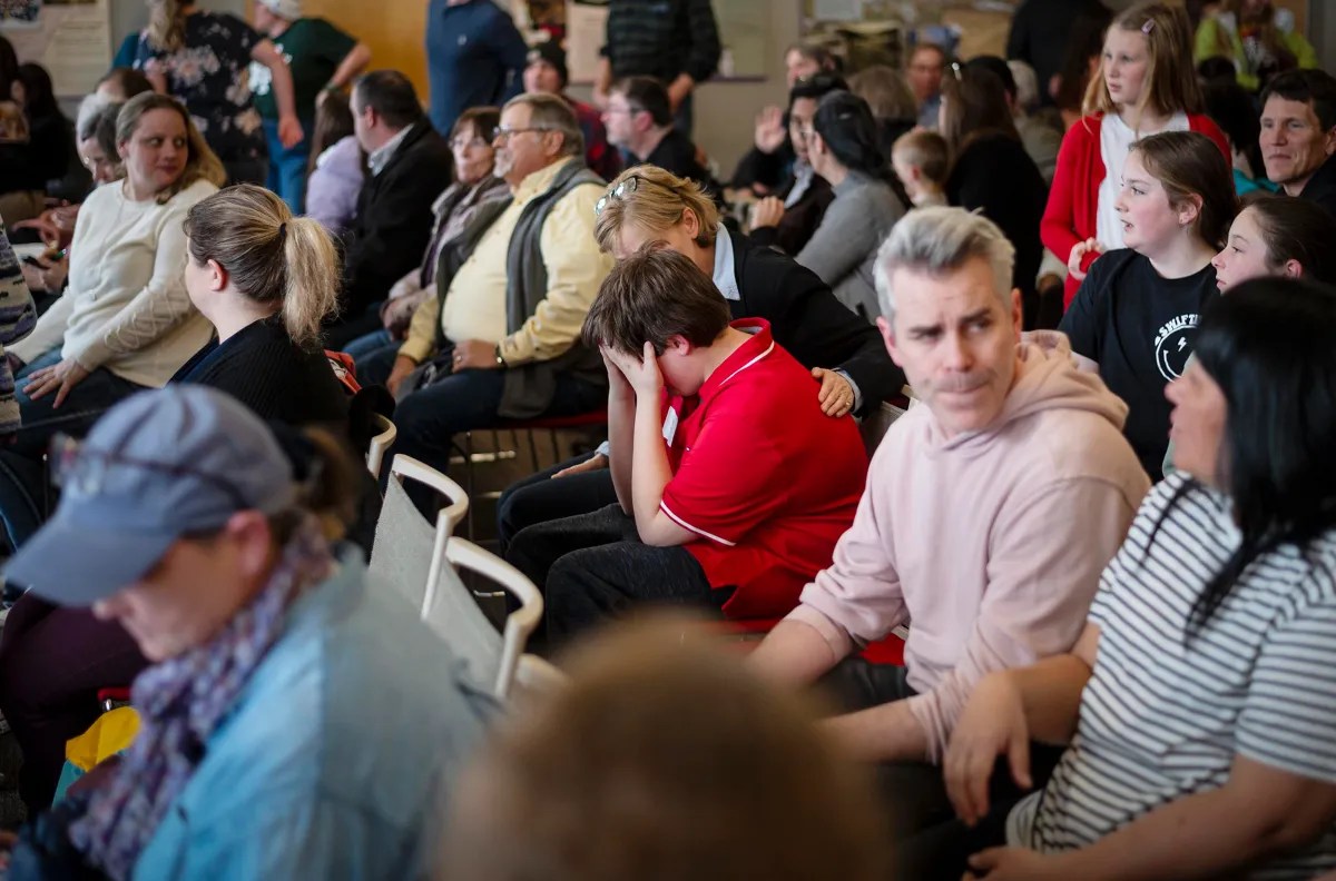 A crowded waiting room with people sitting on chairs, some engaged in conversation while one individual appears to be covering their ears.