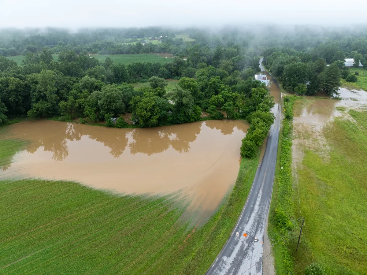 Aerial view of a flooded rural area with submerged fields and a road partially covered by water in a foggy landscape. Trees and green vegetation surround the flooded sections. Traffic cones block the road.