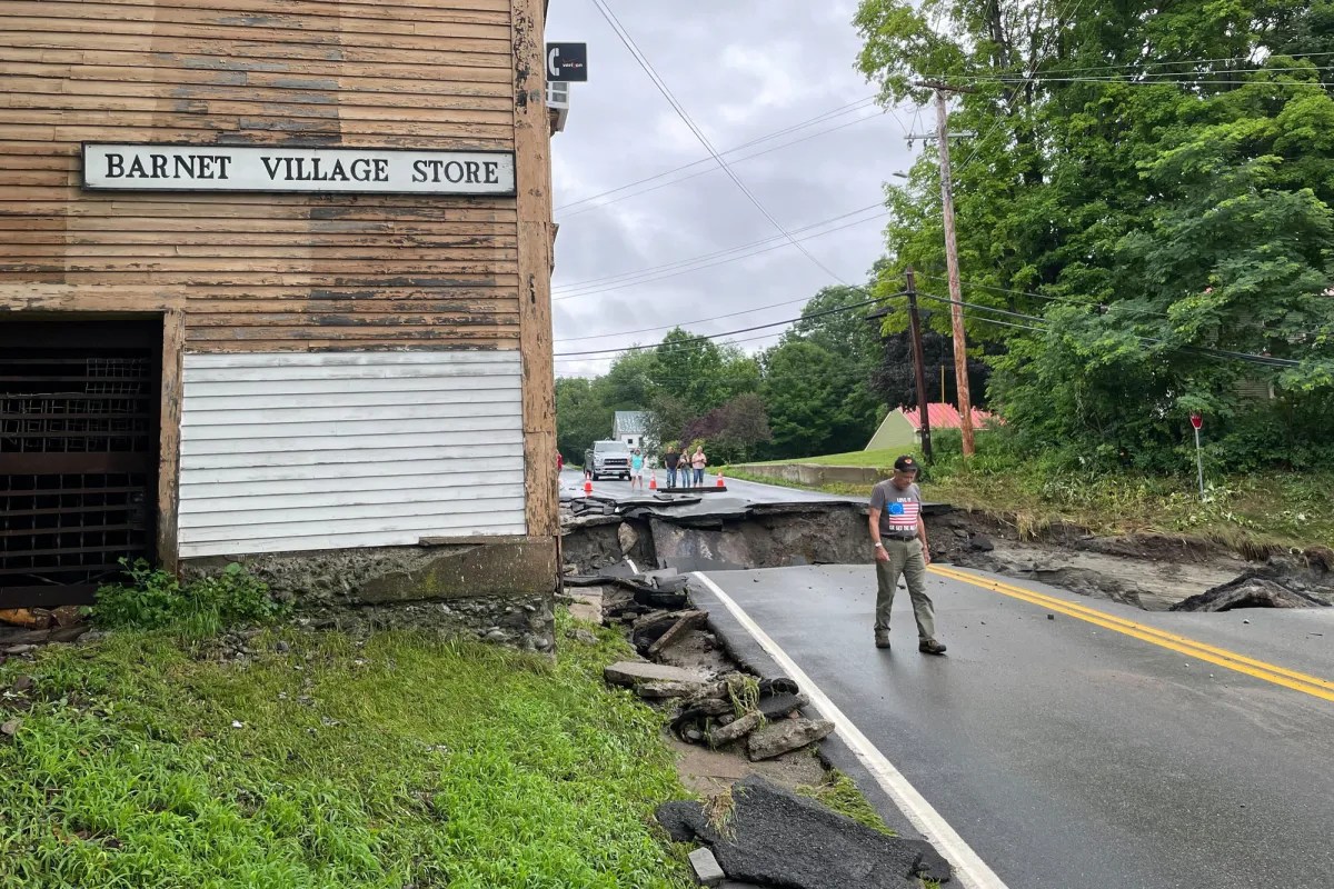 A person stands near a cracked, sunken road next to a Barnet Village Store building. The road is heavily damaged and partially collapsed, with a vehicle nearby. Trees and utility wires are visible.