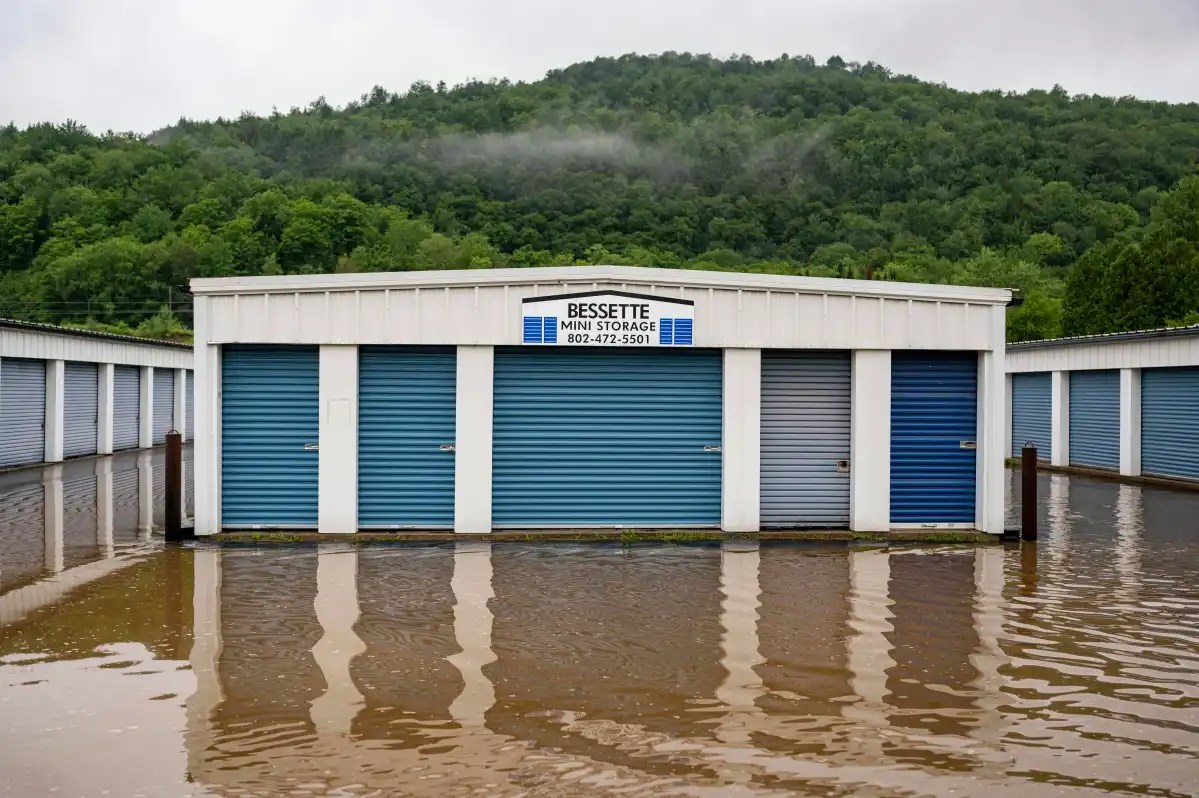 Flooded storage facility with blue and white units, submerged up to their doors. A sign reading "Besette Mini Storage" is visible above the central unit. Trees and hills are in the background.