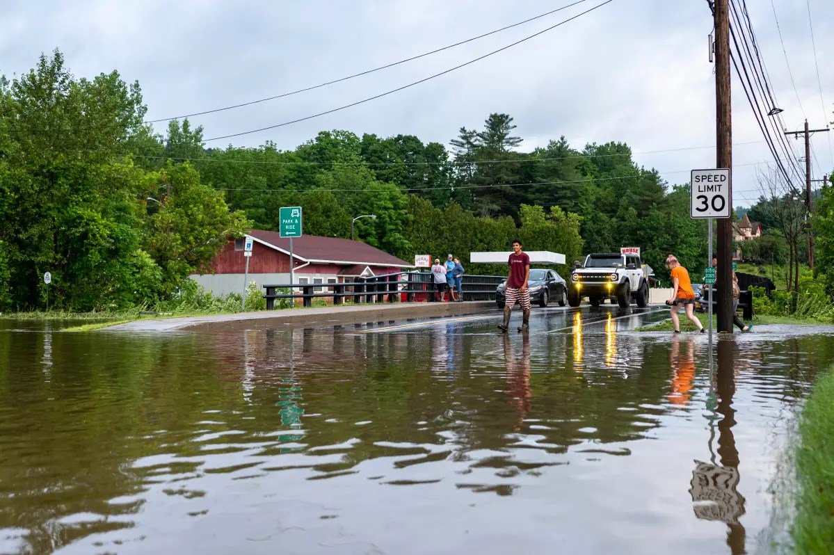 People walking and vehicles halted on a flooded roadway near a 30 mph speed limit sign, with surrounding trees and buildings partially visible.