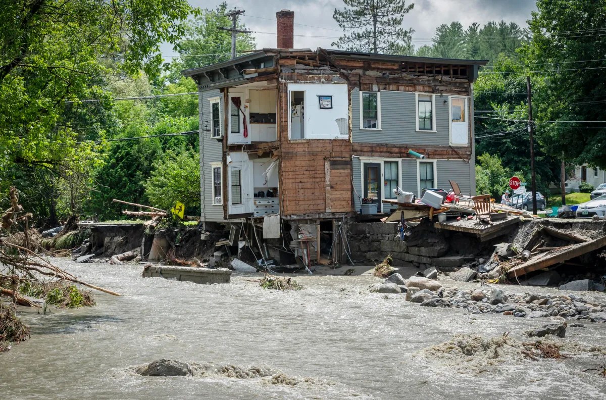 A damaged house stands precariously over a river with missing walls and debris scattered around after a flood. Trees and other houses are visible in the background.